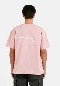 Camiseta de algodón rosa claro con un gran logotipo blanco impreso y texto en la parte posterior, con mangas cortas y un ajuste relajado.