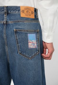 Sinised teksad, millel on beež nahkmärgistus "Fiorucci" kirjatüübiga. Paremas taga taskus on värvikas tikand mägemajadest.