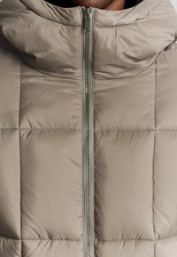 Beige gewatteerde jas met een hoge kraag, voorzien van een centrale rits en een vierkante gestikte drukkleur. Glad, lichtgewicht materiaal met een subtiele glans.