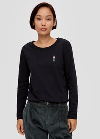 Czarny, długi rękaw top z okrągłym dekoltem, na którym znajduje się mała haftowana postać na piersi; noszony z ciemnymi spodniami z kordurowej tkaniny.