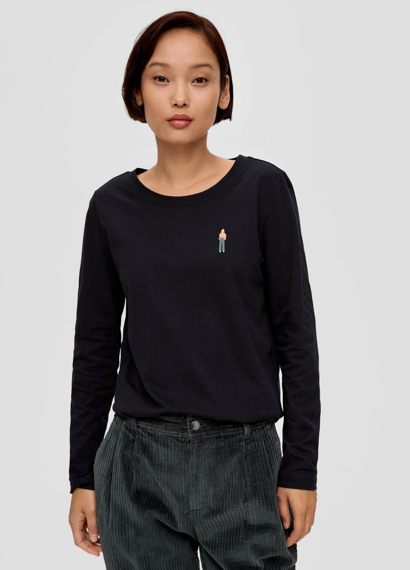 Czarny, długi rękaw top z okrągłym dekoltem, na którym znajduje się mała haftowana postać na piersi; noszony z ciemnymi spodniami z kordurowej tkaniny.