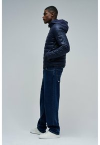 Granatowa kurtka puchowa z kapturem, pikowany design i boczne kieszenie, zestawiona z luźnymi ciemnymi dżinsami i białymi sneakersami.