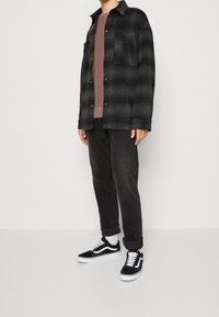 Camicia in flanella a quadri nera e grigia, abbottonata con una tasca sul petto, indossata sopra una T-shirt color malva, abbinata a jeans neri e scarpe Vans nere.