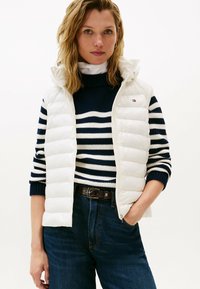 Smanicato imbottito bianco con collo alto, indossato sopra un maglione a righe navy e bianco. Jeans in denim scuro e cintura nera completano l'outfit.