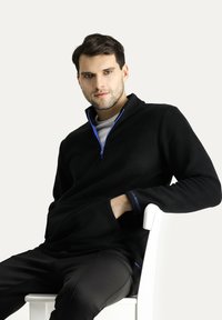 Sudadera de forro polar negra con medio cierre y acentos azules, que presenta un bolsillo frontal y mangas largas. Llevada con pantalones negros frente a un fondo sencillo.