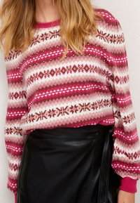 Pull en tricot avec un motif géométrique rose et blanc, agrémenté de accents marron et de embellissements, à manches longues et avec un col côtelé.