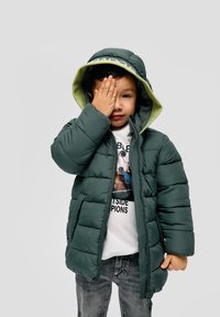 Donkergroene pufferjack met een capuchon, voorzien van een lichtgroene voering, gedragen over een witte graphic t-shirt en grijze jeans.
