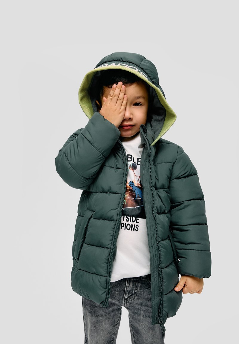 Donkergroene pufferjack met een capuchon, voorzien van een lichtgroene voering, gedragen over een witte graphic t-shirt en grijze jeans.