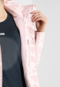Personne portant une chemise noire à motif de points blancs tient ouverte une veste camo rose montrant une poche intérieure zippée et un col.