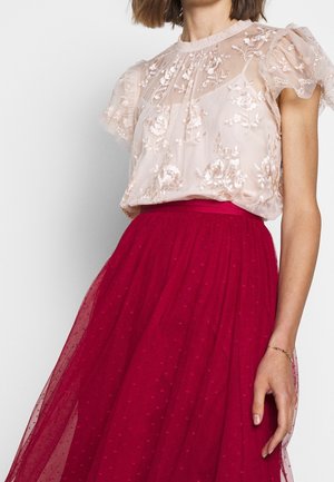Woman wearing a sheer, beige floral embroidered blouse with puff sleeves and a high-waisted, red tulle skirt with small dot patterns.