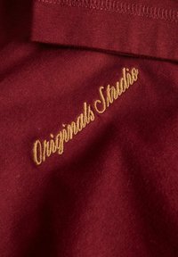 Burgundia kangas, millel on kullatud tikandiga tekst "Originals Studio". Tekstuur on pehme ja sile viimistlus.