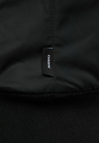 Zwart stof met gestikte naden en een klein zwart label met de tekst "CHASIN'" in witte letters bevestigd aan de naad.