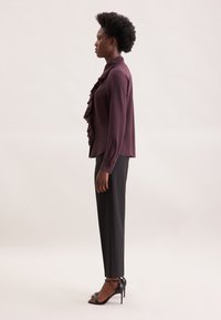Bordeauxfarbige Bluse mit Rüschendetails, langen Ärmeln und spitzem Kragen, kombiniert mit schwarzen, taillierten Hosen und offenen, hohen Absätzen.