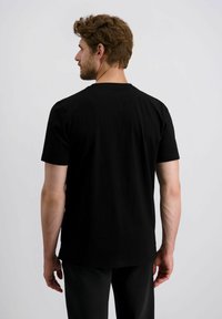 Mann mit braunen Haaren trägt ein schlichtes schwarzes T-Shirt mit kurzen Ärmeln und schwarze Hosen, steht mit dem Rücken zu einem hellgrauen Hintergrund.