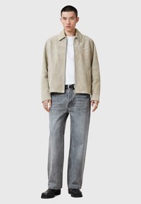Beige mockaskinnjacka med krage, dragkedjestängning och två sidofickor, i kombination med ljusgrå vida jeans och svart skor.