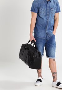 Borsa da viaggio in pelle nera con doppi manici, texture liscia e design minimalista, abbinata a una camicia in denim e pantaloni corti.