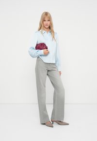 Žena stoji u svijetloplavoj bluzi, siva široka traperica, sive štikle, drži bordo clutch torbu na bijeloj pozadini.