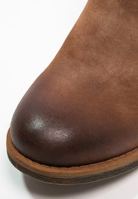 Bout de botte en daim marron avec trépointe cousue et bord de semelle en bois, montrant la texture et un dégradé d'ombrage sur la surface du cuir.