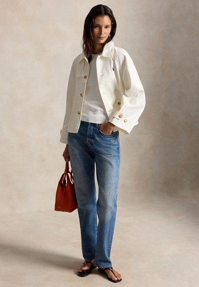 Γυναίκα με λευκό μπουφάν, λευκό πουκάμισο, μπλε τζιν, μαύρα σανδάλια, κρατώντας κόκκινη τσάντα, στέκεται μπροστά σε απλό μπεζ φόντο.