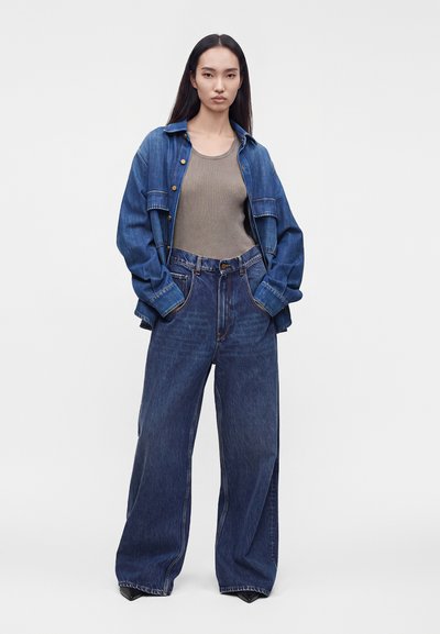 Jeune femme portant un jean en denim bleu foncé ample, un débardeur côtelé beige et une veste en denim bleue déboutonnée, mains dans les poches.