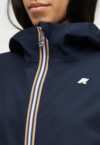 Γυναίκα που φοράει μπλε σκούρο σακάκι με πολύχρωμη ρίγα στον φερμουάρ και λευκό λογότυπο στο στήθος, ο φερμουάρ είναι μερικώς κλειστός κοντά στο πηγούνι.