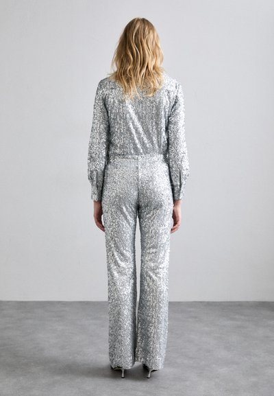 Sølv paljett jumpsuit med lange ermer, tettsittende midje og utsvingte ben. Har en glatt tekstur og høy hals.