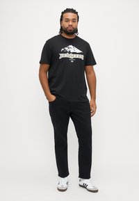 Μαύρο T-shirt με γραφικό βουνό και κείμενο "jack & jones", συνδυασμένο με σκούρο παντελόνι και λευκά αθλητικά παπούτσια με μαύρες λεπτομέρειες.