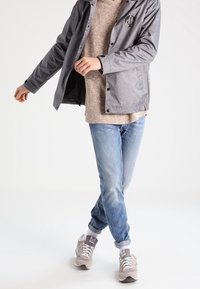 Šedá lehká bunda s černým logem, béžový texturovaný svetr, modré džíny a světle šedé tenisky s bílými tkaničkami. Neformální outfit.