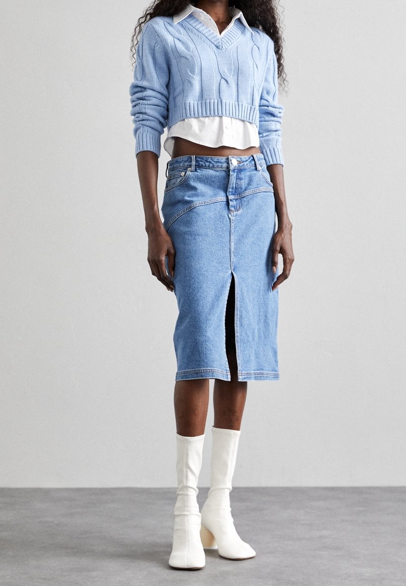 Femme portant un pull court à torsades bleu clair, une chemise blanche, une jupe en jean taille haute avec une fente frontale, et des bottes blanches mi-mollet.