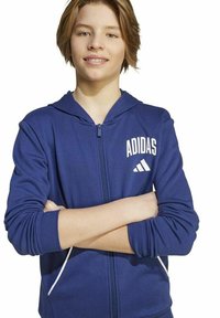 Jongere met kort bruin haar, gekleed in een blauwe Adidas hoodie met rits, staand met gekruiste armen tegen een witte achtergrond.