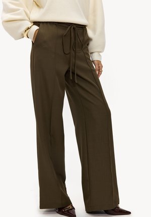 Femme portant un pantalon large vert olive avec un nœud sur le devant, un pull crème, un bracelet en or et des chaussures bordeaux à bout pointu.