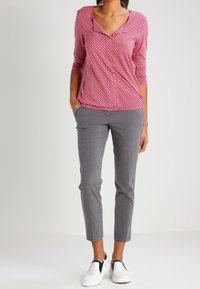 Rood en wit gestreepte lange mouw blouse met V-hals, gecombineerd met grijze slim-fit broek en witte instapschoenen met zwarte accenten.