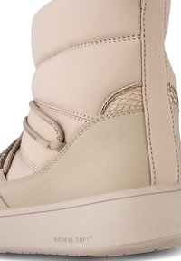 Ljust beige mjukt läderstövel med texturerade accenter, elastiska snören och en kraftig sula märkt "NATURAL SOFT." Har en slät, rundad design.