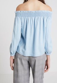 Jasnoniebieska bluzka odsłaniająca ramiona z marszczonym dekoltem, długimi bufkowymi rękawami i miękką bawełnianą tkaniną. Noszona z szarymi, w kratę spodniami.