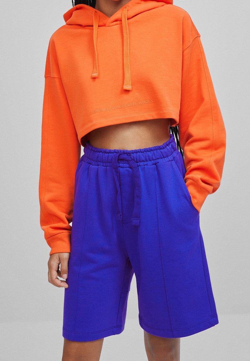Avskuren orange huvtröja med ett framsidestryck och lila shorts med elastisk midja och sidofickor, tillverkade av mjukt tyg.