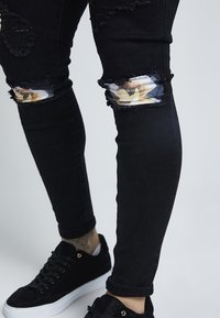 Black, ripped jeans featuring transparent patches with floral print on the knees, fitted design, and standard denim texture. Black sneakers included.