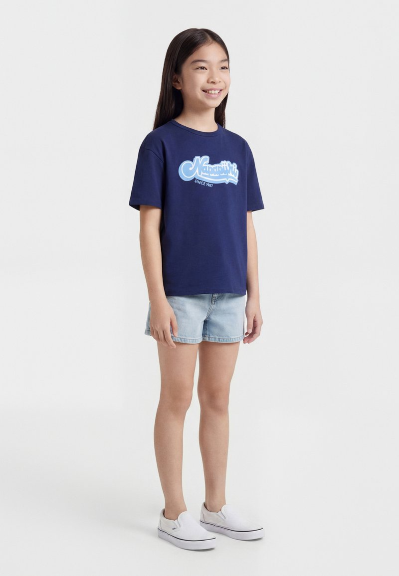 Νεαρή κοπέλα όρθια, φοράει μπλε ναυτικό T-shirt με σχέδιο, ανοιχτόχρωμο τζιν σορτς και λευκά αθλητικά παπούτσια χωρίς κορδόνια, χαμογελά και κοιτάζει ελαφρώς δεξιά.