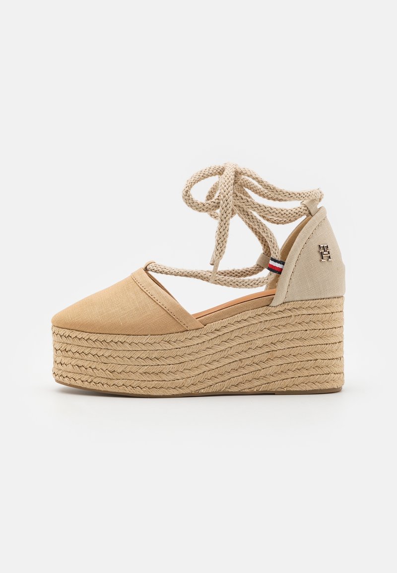 Espadrille compensée beige avec semelle en corde tressée, liens à nouer autour de la cheville, bout fermé et petit logo sur le contrefort du talon.