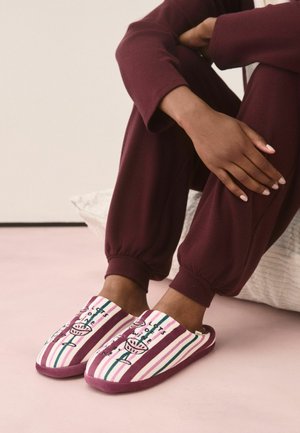 Person trägt bordeauxrote Hose und gestreifte rosa-weiße Hausschuhe mit Cocktailglas und Textdesign und sitzt auf dem Boden.