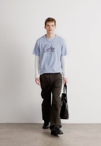 Jasnoniebieska koszulka z krótkim rękawem z grafiką "Calvin", założona na białą koszulę z długim rękawem. Brązowe spodnie o szerokich nogawkach i czarne buty. Czarna torba.