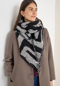 Svart och grå scarf med geometriskt mönster, gjord av texturerad tyg, med fransar i kanterna, bärs över en brun kappa.