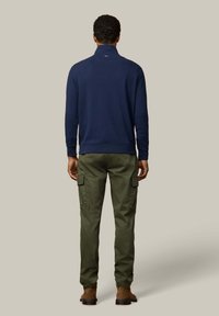 Pullover blu navy con mezza cerniera e pantaloni cargo color oliva. Il pullover presenta un collo alto; i pantaloni includono tasche cargo laterali e gambe affusolate. Scarpe marroni.