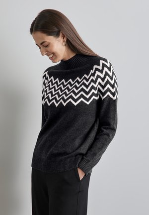 Pull noir avec un motif en zigzag blanc sur la poitrine, présentant un col haut et des poignets côtelés. Associé à un pantalon noir.