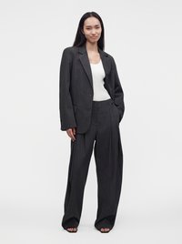 Γυναίκα με μαύρο oversized κοστούμι με λεπτές ρίγες και φαρδιά παντελόνια, καθώς και λευκό αμάνικο τοπ, στέκεται με το ένα χέρι στην τσέπη.