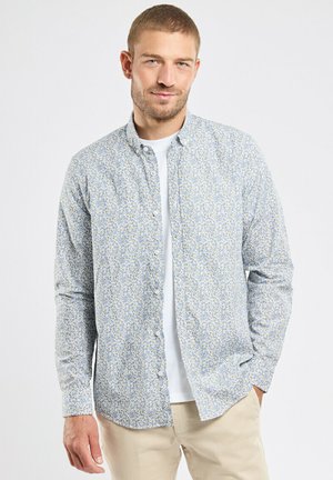 Homme portant une chemise à boutons bleu clair à motif floral sur un t-shirt blanc, pantalon beige, debout avec une main dans la poche devant un arrière-plan uni.