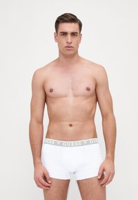 Boxerii albi cu talie elastică gri, având logo-ul "GUESS". Materialul pare moale, cu un aspect ușor texturat.