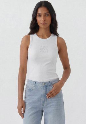 Frau mit langen dunklen Haaren, die ein weißes ärmelloses Oberteil mit dem Schriftzug „sun love oui“ und hellblaue High-Waist-Jeans trägt, steht vor weißem Hintergrund.