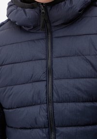 Giubbotto imbottito di colore blu navy scuro con motivo a righe orizzontali, colletto alto, zip frontale completa e tessuto leggero e testurizzato.