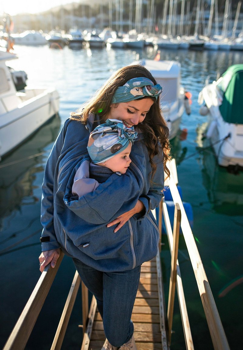 Zweischichtige blaue Softshelljacke mit Kapuze für ein Kind. Gemusterter blauer Schal. Holzsteg in einer Marina mit Booten im Hintergrund.