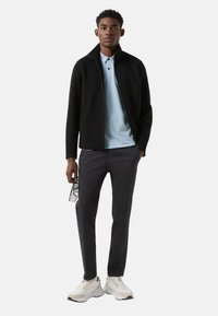 Zwarte jas met ritssluiting, lichtblauw poloshirt, donkere broek en beige sneakers. Model draagt een bril; casual, gelaagde look.